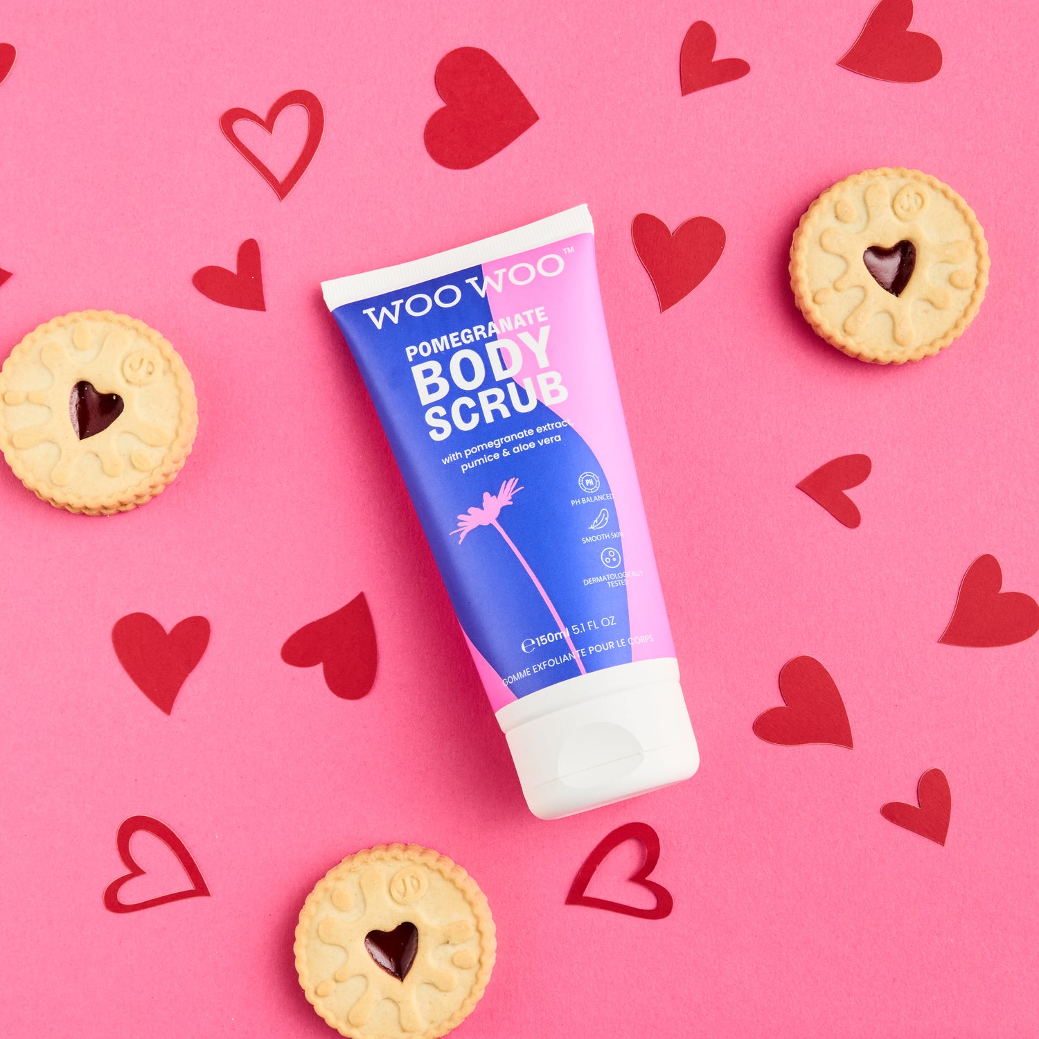 Woo Woo Pomegranate Body Scrub tube on a pink background with heart shapes and cookies.